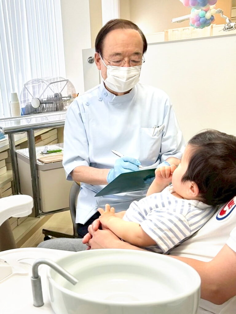 診療台に親御さんと一緒に座るお子様とカルテに記入している様子の鯨井院長の写真